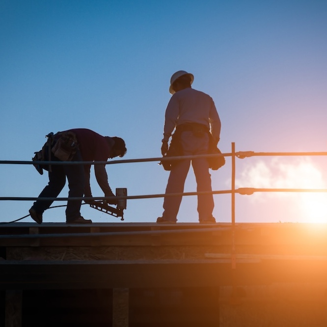 Two roofers are silouetted by a setting sun on a construction site.